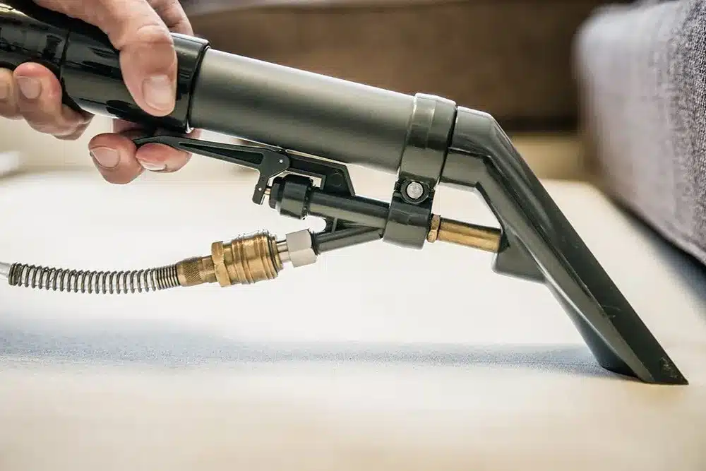 Close-up of a hand operating a professional carpet cleaning wand, focusing on the clean spray extraction on a light-colored carpet