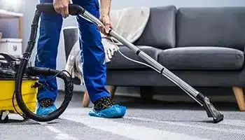 A person in blue uniform and shoe covers vacuuming a carpet with a professional-grade vacuum cleaner, indicating a cleaning service in progress