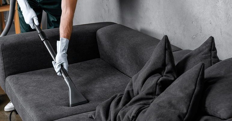 Professional cleaning service employee wearing gloves vacuuming a dark gray sofa with a handheld upholstery cleaner.